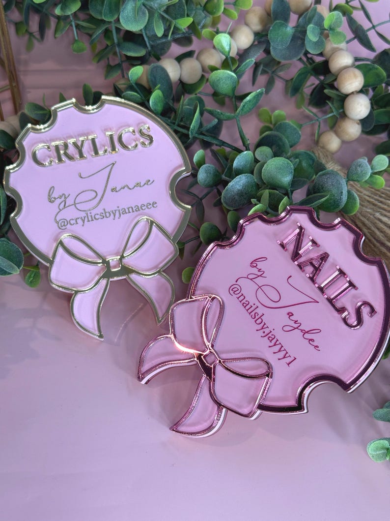 May include: Two pink and gold acrylic signs with bow accents. One sign reads "CRYILCS by Janae @crylicsbyjanaeee", the other reads "NAILS by Jaydee @nailsby.jayyy1". The signs are surrounded by greenery.