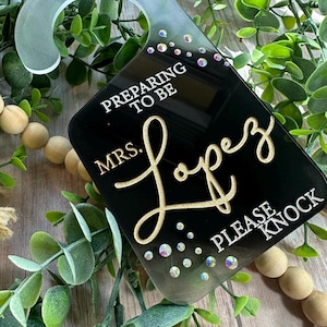 Könnte beinhalten: Schwarzer Türhänger mit dem Text "PREPARING TO BE MRS. Lopez PLEASE KNOCK" in goldener und weißer Schrift. Der Aufhänger hat eine transparente Oberseite mit schillernden Strasssteinen und einen gebogenen Griff.