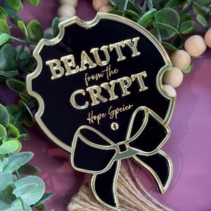 May include: A black and gold decorative pin with the text "BEAUTY from the CRYPT" and a bow design. The pin has a gold border and the text is in gold. The pin is surrounded by greenery and wooden beads.
