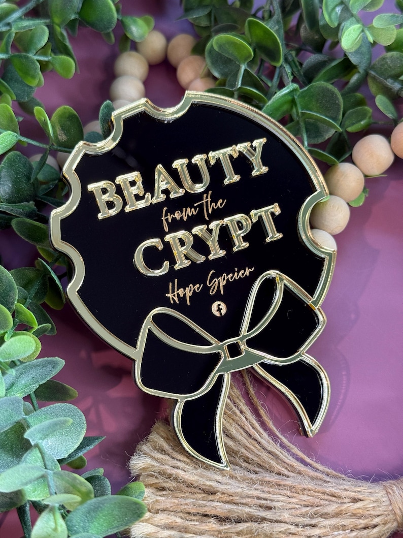 May include: A black and gold decorative pin with the text "BEAUTY from the CRYPT" and a bow design. The pin has a gold border and the text is in gold. The pin is surrounded by greenery and wooden beads.