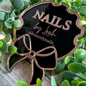 May include: A black and rose gold sign with the text "NAILS by Leah" and the Instagram handle "@leisurelynails_". The sign has a decorative bow and is surrounded by greenery. The sign is likely used for nail salon promotion.