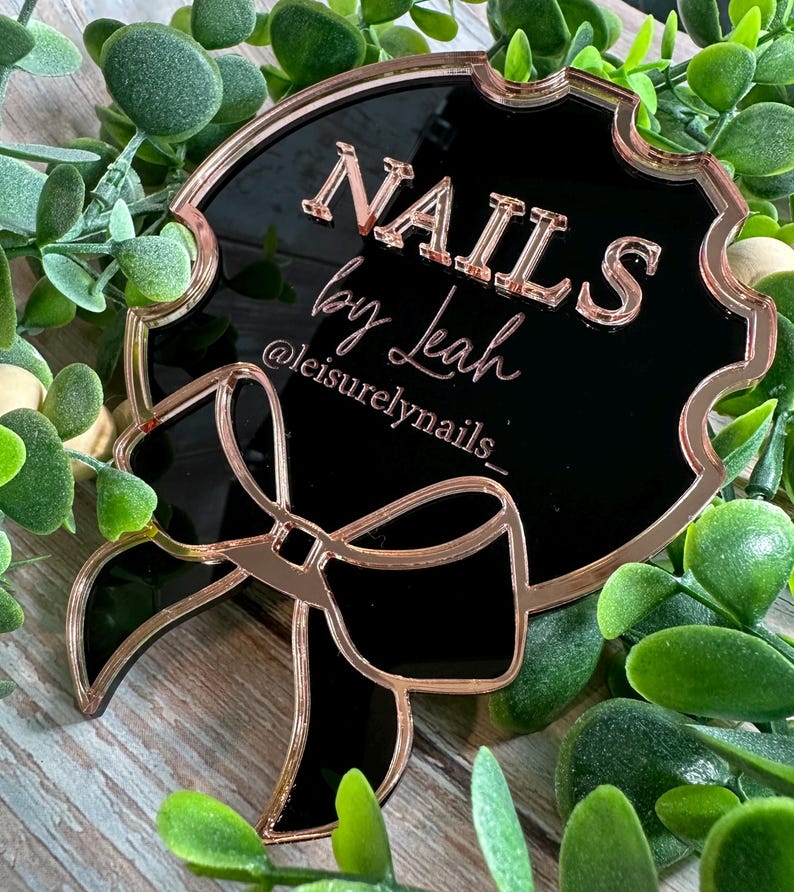 May include: A black and rose gold sign with the text "NAILS by Leah" and the Instagram handle "@leisurelynails_". The sign has a decorative bow and is surrounded by greenery. The sign is likely used for nail salon promotion.