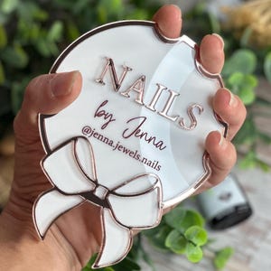 May include: A white and rose gold acrylic sign with the text "NAILS by Jenna" and the Instagram handle @jenna.jewels.nails. The sign has a bow design and is held by a hand.