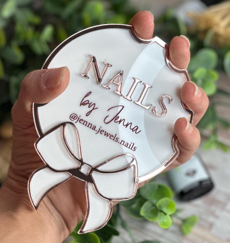 May include: A white and rose gold acrylic sign with the text "NAILS by Jenna" and the Instagram handle @jenna.jewels.nails. The sign has a bow design and is held by a hand.