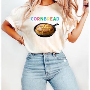 May include: Cream-colored t-shirt with the word "CORNBREAD" in rainbow letters above an image of cornbread in a cast iron skillet. The shirt is paired with light blue jeans.
