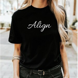 Può includere: T-shirt nero con la scritta "Align" in corsivo bianco. La maglietta è a girocollo e sembra essere realizzata in un materiale morbido. La modella indossa jeans neri e un braccialetto.