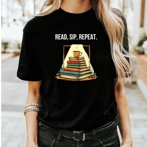 以下が含まれることがあります： 「READ, SIP, REPEAT」の文字が入った黒いTシャツ。本が積み重ねられ、その上にコーヒーカップが置かれたグラフィックが特徴です。本は様々な色で描かれ、デザインは薄い黄色の枠で囲まれています。