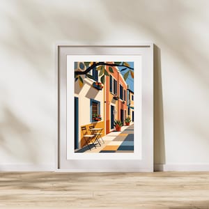 May include: A framed art print depicting a street scene with orange and cream-coloured buildings, a small table with two chairs, potted plants, and tree branches overhead. The illustration features a geometric pavement pattern in blue, yellow, and grey.