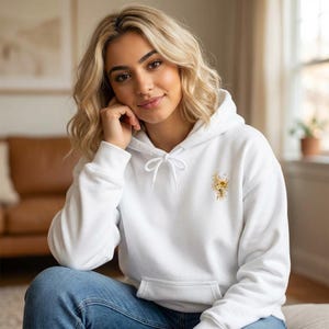 Peut inclure: Un sweat à capuche blanc avec un petit motif floral sur la poitrine. Le sweat à capuche a une capuche à cordon et une poche kangourou. Le motif floral présente des tournesols et d'autres éléments jaunes et dorés. Le modèle porte un jean bleu.
