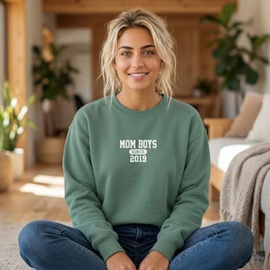 Peut inclure: Un sweat-shirt à col rond vert sauge avec une inscription blanche imprimée sur la poitrine indiquant MOM BOYS dans une police sans empattement, avec le mot SINCE et l'année 2019 en plus petit juste en dessous du texte principal.