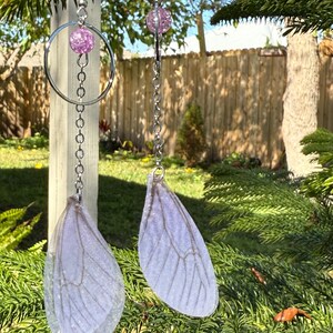 May include: A pair of dangle earrings with a silver chain and a clear, iridescent, purple, glitter-filled bead. The earrings feature a clear, iridescent, purple, glitter-filled bead at the top and a clear, iridescent, purple, glitter-filled wing charm at the bottom.