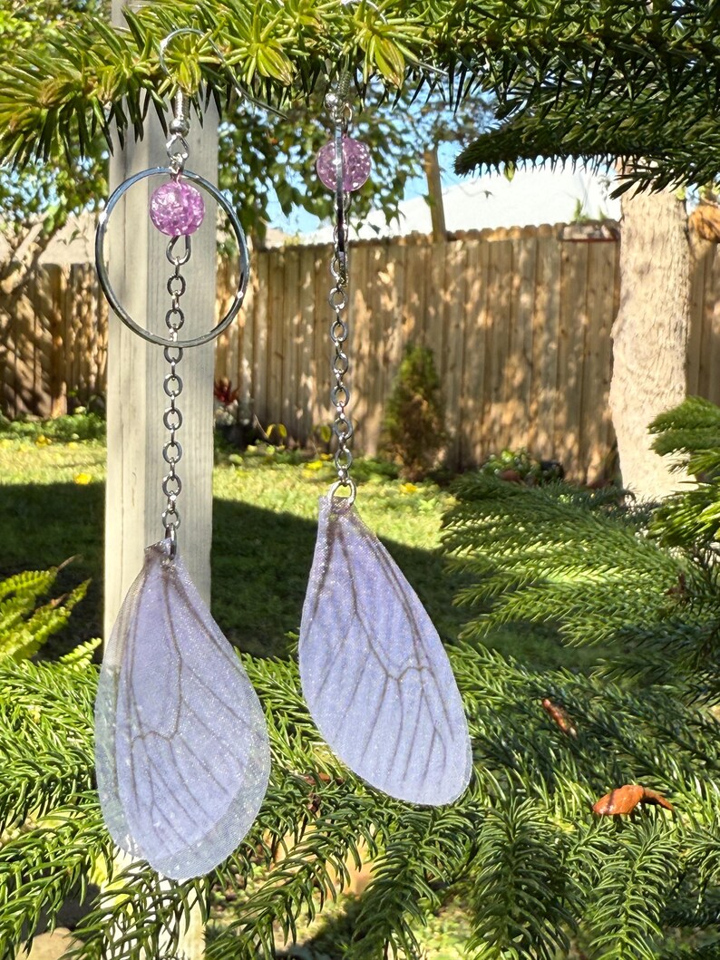 May include: A pair of dangle earrings with a silver chain and a clear, iridescent, purple, glitter-filled bead. The earrings feature a clear, iridescent, purple, glitter-filled bead at the top and a clear, iridescent, purple, glitter-filled wing charm at the bottom.