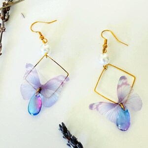 May include: A pair of delicate butterfly earrings. Each earring features a gold-colored square frame, a white pearl, and a translucent lavender butterfly with a teardrop-shaped iridescent charm. The earrings are designed with a hook closure.