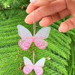 May include: Two butterfly-shaped earrings with pink and white wings are displayed against a backdrop of green foliage. The earrings are suspended from thin silver wires. The butterfly wings have a delicate, translucent appearance.