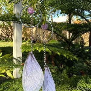 May include: A pair of silver hoop earrings with a dangling chain and a purple iridescent bead. The chain ends with a white, iridescent, translucent wing-shaped charm.
