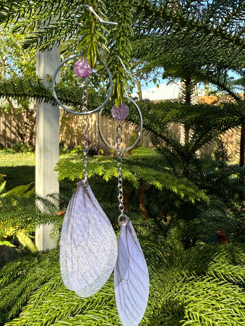 May include: A pair of silver hoop earrings with a dangling chain and a purple iridescent bead. The chain ends with a white, iridescent, translucent wing-shaped charm.