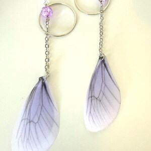 May include: A pair of silver earrings with a delicate chain and a purple bead. Each earring features a translucent white wing with black veins, resembling a dragonfly wing.