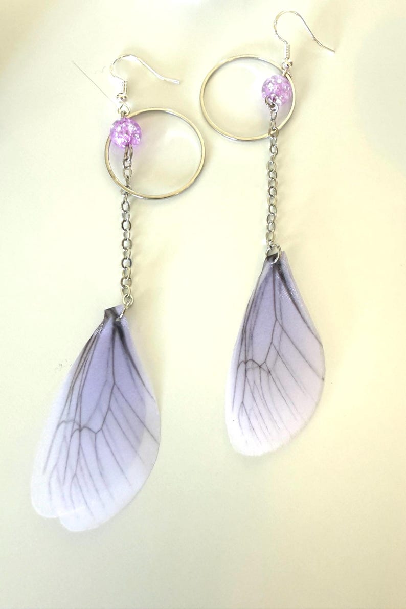 May include: A pair of silver earrings with a delicate chain and a purple bead. Each earring features a translucent white wing with black veins, resembling a dragonfly wing.