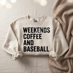 May include: Cream-colored sweatshirt with the words "WEEKENDS COFFEE AND BASEBALL" printed in bold black letters. The sweatshirt has a crew neck and long sleeves with cuffed wrists. The background is a neutral color.