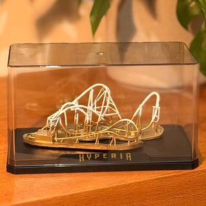 Puede incluir: Maqueta dorada de montaña rusa dentro de una vitrina transparente. El modelo presenta un diseño detallado de la pista con soportes blancos. La base negra tiene la palabra "HYPERIA" en blanco. La vitrina está sobre una superficie de madera.