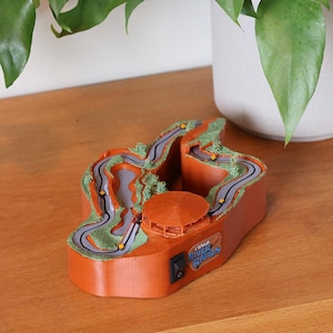 May include: A 3D printed model of the Congo River Rapids ride featuring a winding grey track, green textured landscape, and a central orange gazebo. The base is orange with a black power switch and a plaque reading Congo River Rapids.