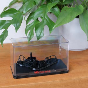 Puede incluir: Un modelo en miniatura negro de una montaña rusa dentro de una vitrina transparente. La base es negra, con el texto "SAW THE RIDE" en rojo. El modelo se encuentra sobre un fondo de hojas verdes.