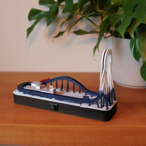 Puede incluir: Modelo en miniatura de montaña rusa con pista azul y soportes blancos. El modelo presenta un bucle y un pequeño vagón de tren. La base es negra con un interruptor de encendido/apagado. El modelo se exhibe sobre una superficie de madera.