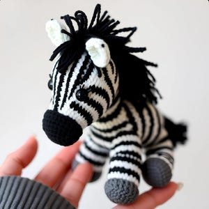 May include: A black and white crocheted zebra soft toy with black eyes and a grey crocheted nose. The zebra is sitting on a hand.