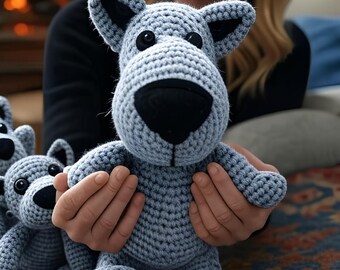 Può includere: Un peluche a forma di cane all'uncinetto blu chiaro con occhi neri e naso nero. Il cane ha grandi orecchie ed è tenuto da una persona. Un altro giocattolo simile è visibile sullo sfondo. I giocattoli sono su un tappeto fantasia.