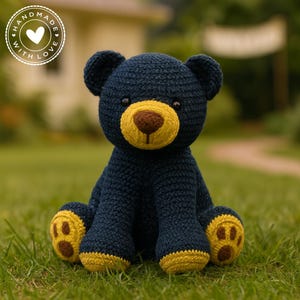 May include: A handmade navy blue crochet bear with yellow and brown accents. The bear has a yellow muzzle and paw pads, and a brown nose. The bear is sitting on green grass. The image includes a circular logo that says "Handmade with Love".