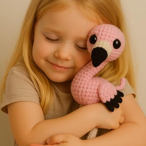 May include: A handmade pink crochet flamingo toy with black detailing on the beak and feet. The flamingo has large, round black eyes. The soft toy is being held by a person.
