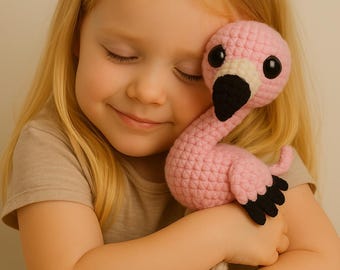 May include: A handmade pink crochet flamingo with black accents, including the beak and feet. The flamingo has large, round black eyes and is being held by a person. The flamingo is a soft toy.