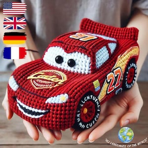 May include: A red crocheted toy car featuring white windshields with black circular eyes, a black mouth, and the number 27 in white and red on the side. The hood displays a gold emblem, and the wheels are black with red centers and white text.