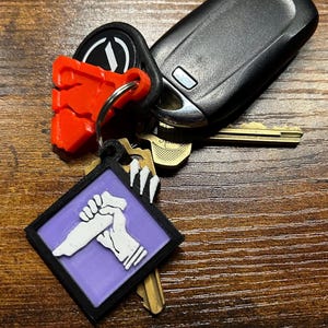 May include: A purple and black keychain with a white graphic of a hand holding a knife. The keychain is attached to a set of keys and a red plastic keychain.
