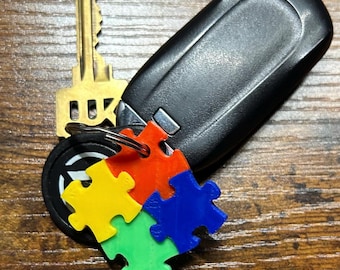 May include: A key chain with a black car key fob and a puzzle piece charm. The puzzle piece charm is made of four pieces, each a different color: yellow, red, blue, and green.