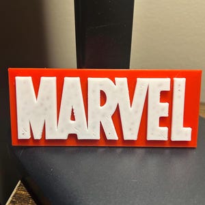 May include: A rectangular sign with the word "MARVEL" in white, raised letters against a bright red background. The sign is likely a decorative item or logo.