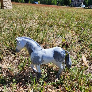 May include: A small, hand-painted horse figurine in a field of grass. The toy horse is white with gray patches and a gray mane and tail. The figurine is standing in a natural outdoor setting.