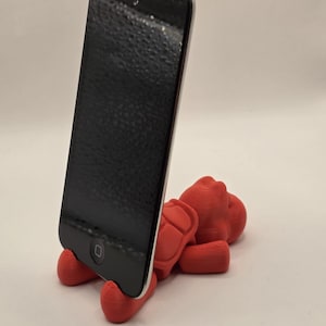 Könnte beinhalten: Ein roter, 3D-gedruckter Telefonständer in Form einer Figur, der ein schwarzes Smartphone hält. Der Ständer hat ein detailliertes Design und stützt das Telefon in aufrechter Position. Der Bildschirm des Telefons ist schwarz.