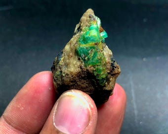 Puede incluir: Una piedra preciosa de esmeralda en bruto, sin cortar, con cristales verdes vibrantes incrustados en una matriz de roca marrón. Los cristales de esmeralda son el punto focal, mostrando su belleza natural y en bruto. La piedra se sostiene en una mano.