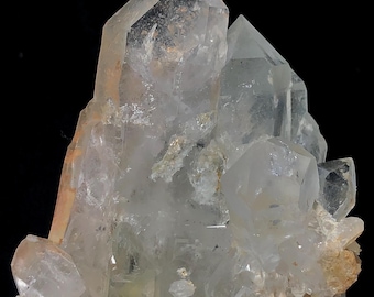 May include: A cluster of clear quartz crystals with sharp, pointed terminations. The crystals have a translucent appearance with subtle internal textures and a light brown hue in some areas. The quartz is mounted on a metal stand against a black background.