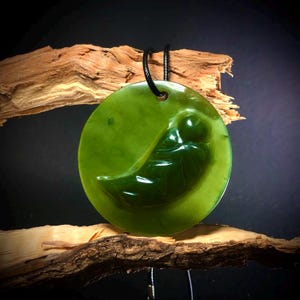 Puede incluir: Un colgante redondo de jade verde con un diseño curvo tallado. El colgante está suspendido de un cordón negro y se muestra sobre un fondo oscuro con un trozo de madera.