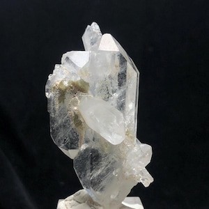 Puede incluir: Un grupo de cristales de cuarzo transparente con múltiples puntas y una base rugosa y texturizada. El cristal es transparente con algunas inclusiones internas y algunas zonas de coloración verde claro y marrón.