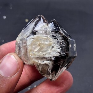 Puede incluir: Un grupo de cristales de cuarzo transparente y ahumado. El cristal tiene una forma geométrica multifacética con una coloración oscura, casi negra, en algunas facetas. El interior tiene una apariencia más clara y translúcida. El cristal se sostiene en una mano.
