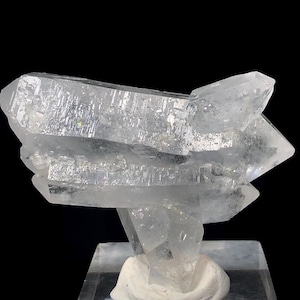 May include: A clear quartz crystal cluster with multiple points. The crystal is mounted on a clear base with a white support. The quartz has a translucent appearance with visible internal structures and a slight iridescent sheen.