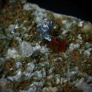 Puede incluir: Primer plano de un espécimen mineral. Presenta estructuras cristalinas transparentes con tonos verdes y marrones. Destacan dos estructuras cúbicas oscuras: una marrón rojizo y otra azul-gris oscuro. La imagen resalta las texturas naturales.