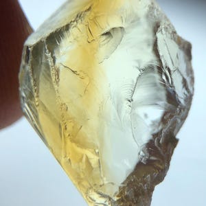 Könnte beinhalten: Ein roher, durchscheinender Edelstein in einem blassgelben Farbton. Der Stein weist ein facettiertes Aussehen mit sichtbaren inneren Rissen und Fehlern auf. Die Kanten sind rau, und die Gesamtform ist unregelmäßig, was auf einen natürlichen, unpolierten Zustand hindeutet.