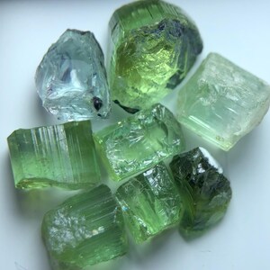 Könnte beinhalten: Eine Sammlung roher, ungeschliffener Edelsteine in Grüntönen und ein blau-grüner Stein. Die Edelsteine haben eine raue, natürliche Textur und unterschiedliche Formen, wobei sich das Licht auf ihren Facetten spiegelt. Die Steine liegen auf weißem Grund.