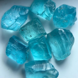 Könnte beinhalten: Eine Sammlung von acht grob geschnittenen, durchscheinenden blau-grünen Edelsteinen. Die Steine haben unregelmäßige Formen und unterschiedliche Größen mit sichtbaren inneren Texturen und Facetten. Die Farbe ist ein lebendiges, klares Aqua.