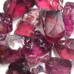 Könnte beinhalten: Nahaufnahme von mehreren grob geschliffenen, tiefrot-violetten Edelsteinen. Die Steine haben unregelmäßige Formen und unterschiedliche Größen mit einem durchscheinenden Aussehen. Die Edelsteine sind zusammengefasst und zeigen ihre natürlichen, unpolierten Oberflächen. Die Farbe reicht von einem dunklen, fast schwärzlichen Rot bis zu einem leuchtenden, helleren Violett.