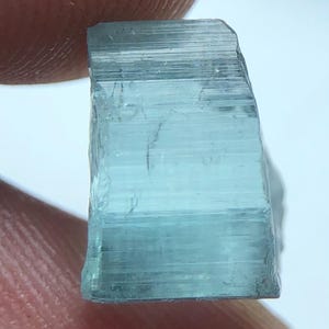 Könnte beinhalten: Nahaufnahme eines hellblauen, rechteckigen Edelsteins, der zwischen Fingern gehalten wird. Der Edelstein hat ein durchscheinendes Aussehen mit sichtbaren linearen Streifen. Die Farbe ist ein blasses, meergrünes Farbschema. Die Kanten des Steins sind rau und uneben.